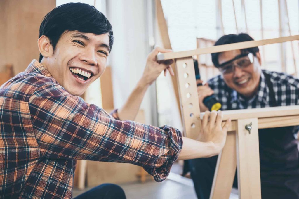 Two people in plaid shirts are smiling and working together on assembling a wooden structure, possibly a piece of furniture, in a bright indoor setting. One person is looking at the camera and laughing.