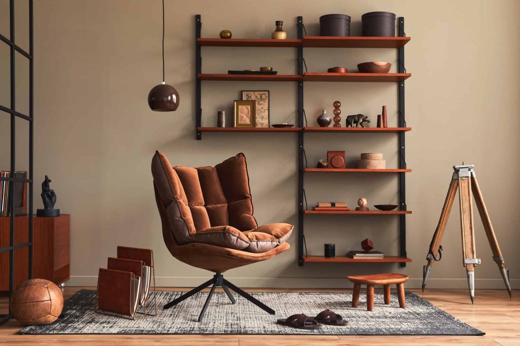 A cozy, modern room with a brown cushioned swivel chair, minimalist shelves holding decor items, a tripod floor lamp, a small wooden side table, and a gray rug on a wooden floor.